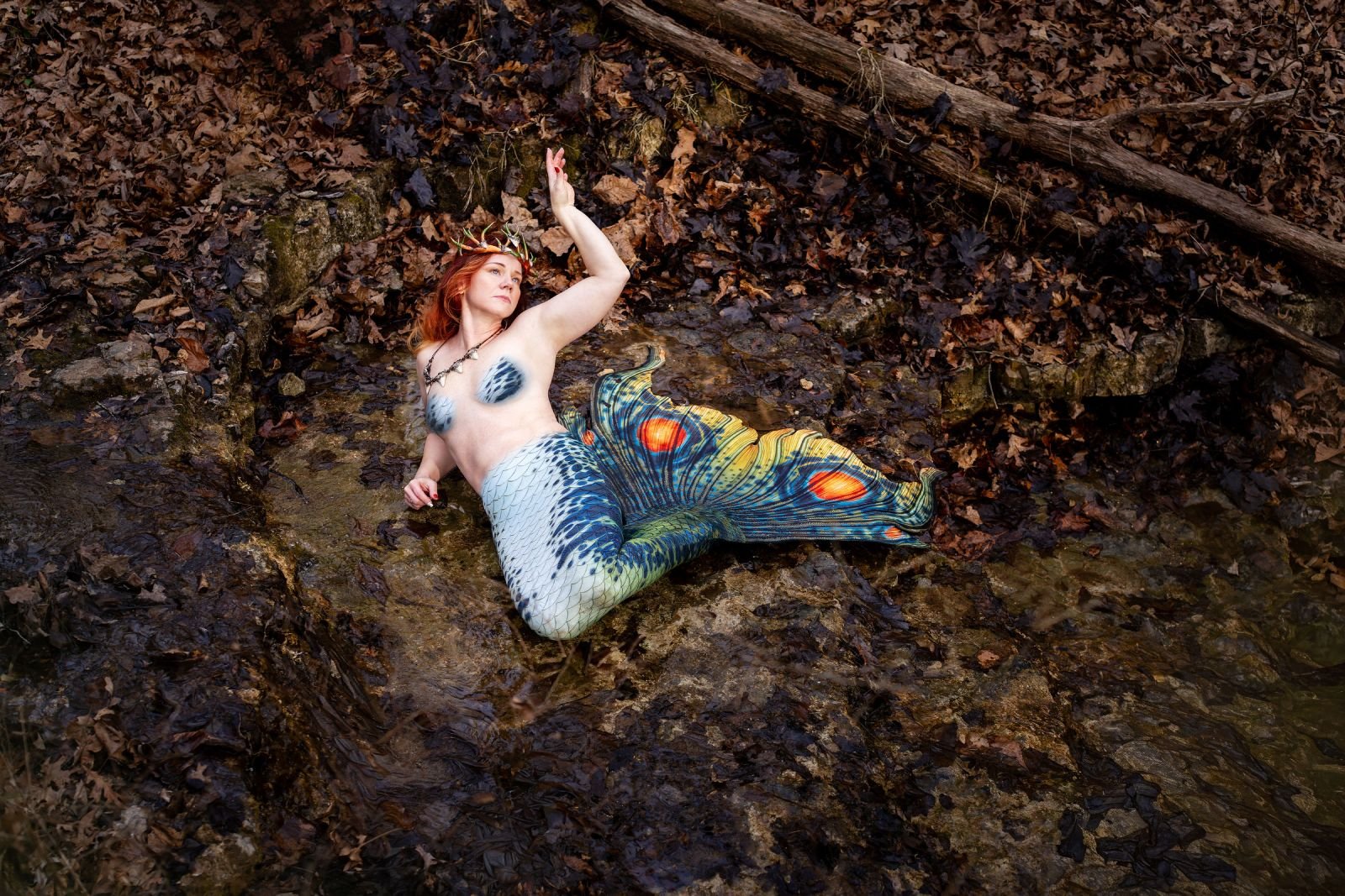 Mermaid performer wearing an antler crown and patterned green tail reclining in a shallow forest creek.