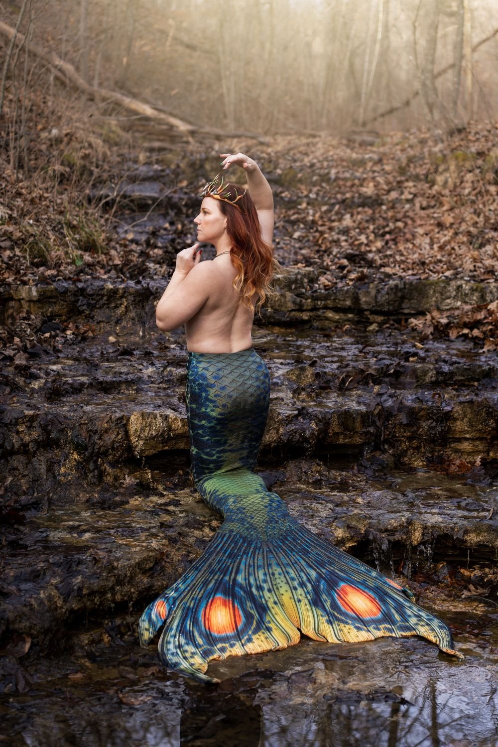 Antler-crowned mermaid performer with a patterned green tail posing in a shallow rocky stream.