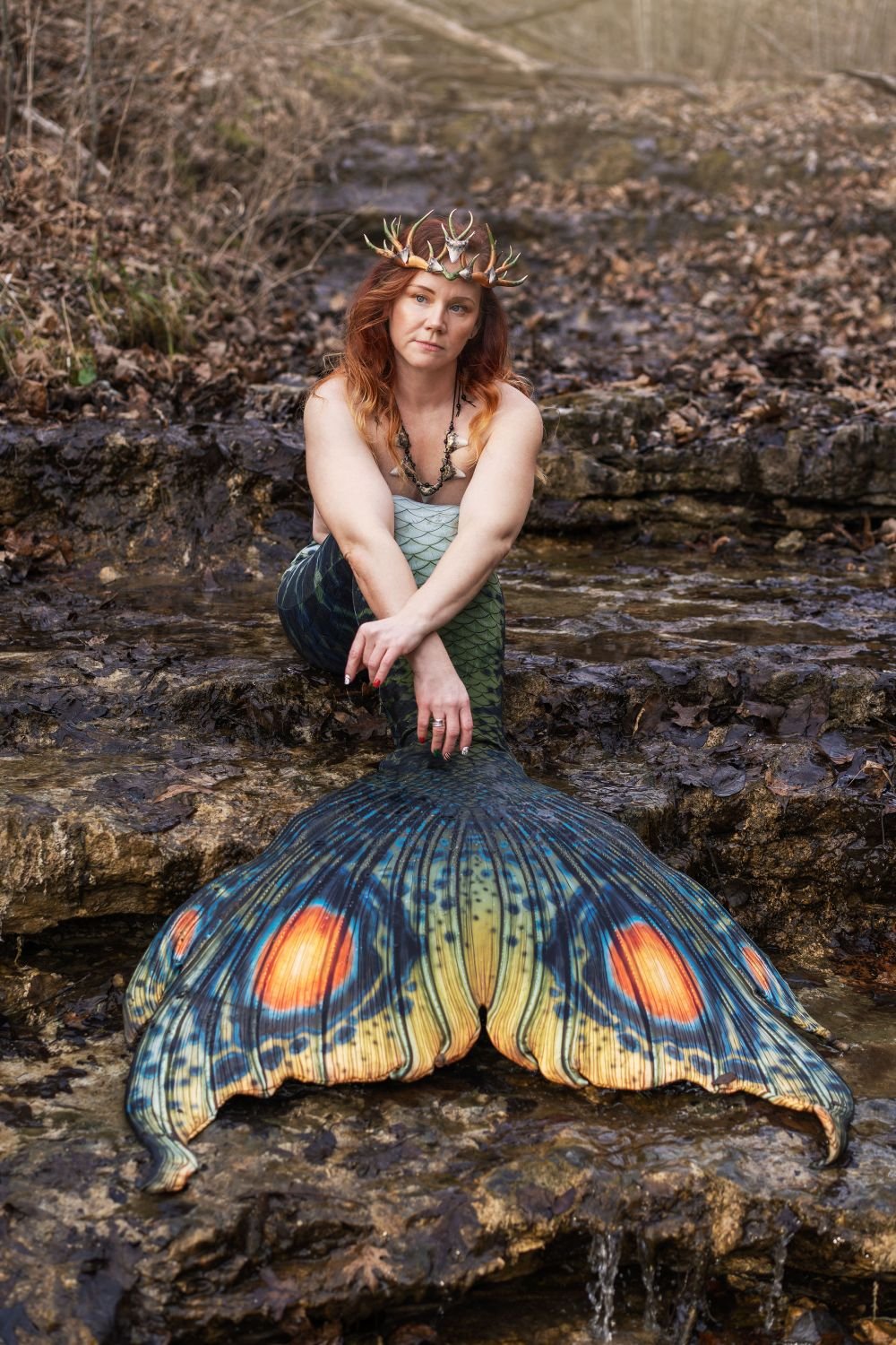 Antler-crowned mermaid performer seated in a rocky woodland stream with a green patterned tail.