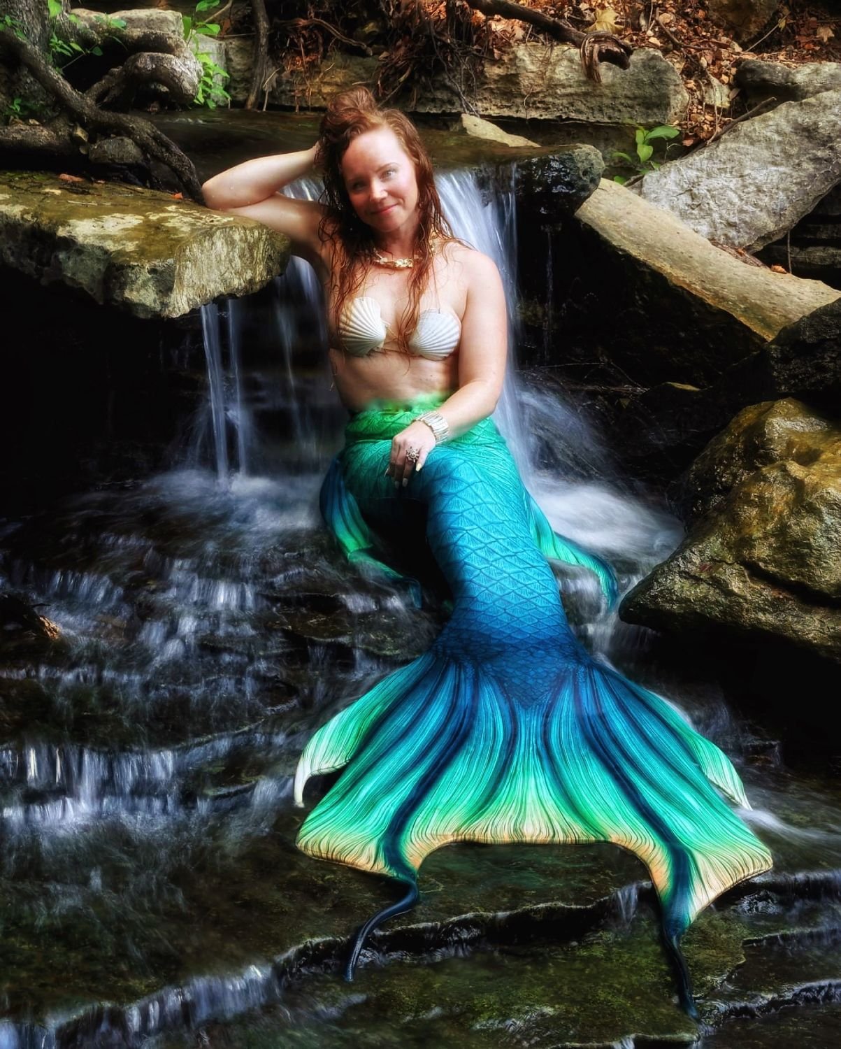 Mermaid performer with a blue-green tail seated beneath a small rocky waterfall.