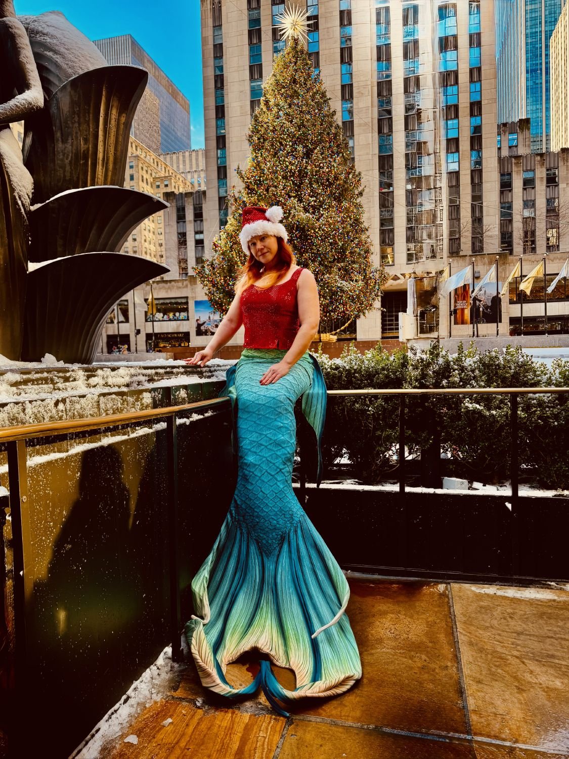 Mermaid performer wearing a Santa hat and turquoise tail beside a decorated Christmas tree.