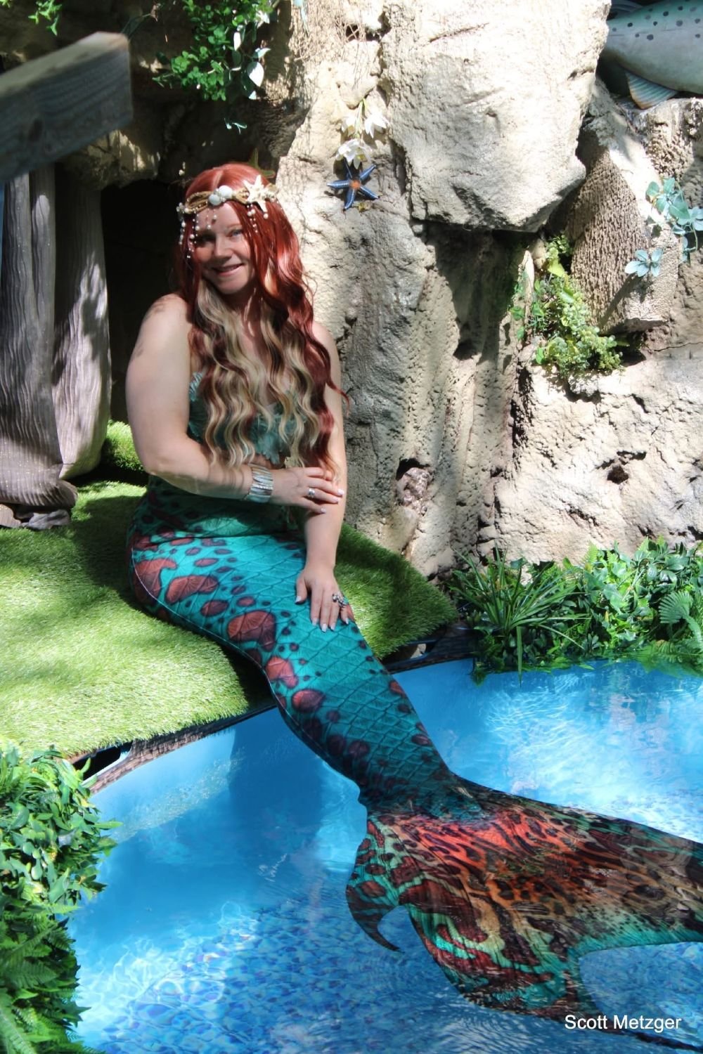 Mermaid performer with a colorful turquoise tail seated beside a decorative blue water display.