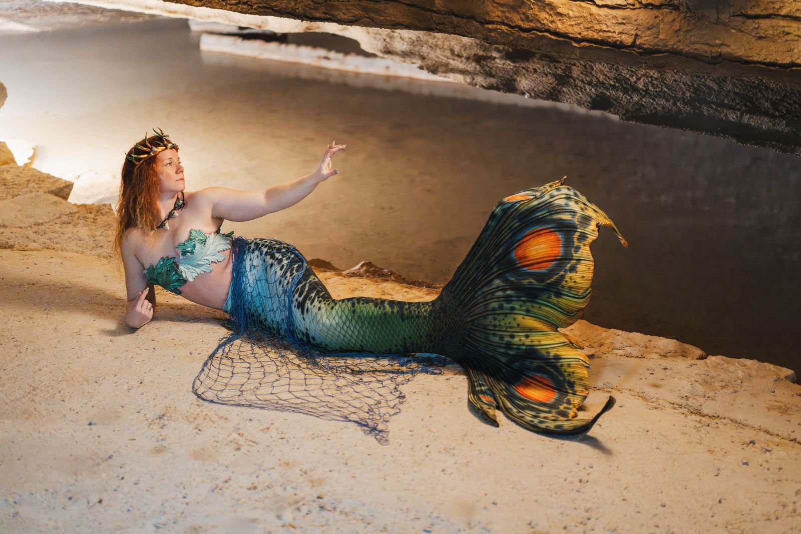 Crowned mermaid performer with a patterned green tail reclining beside a still cave pool.