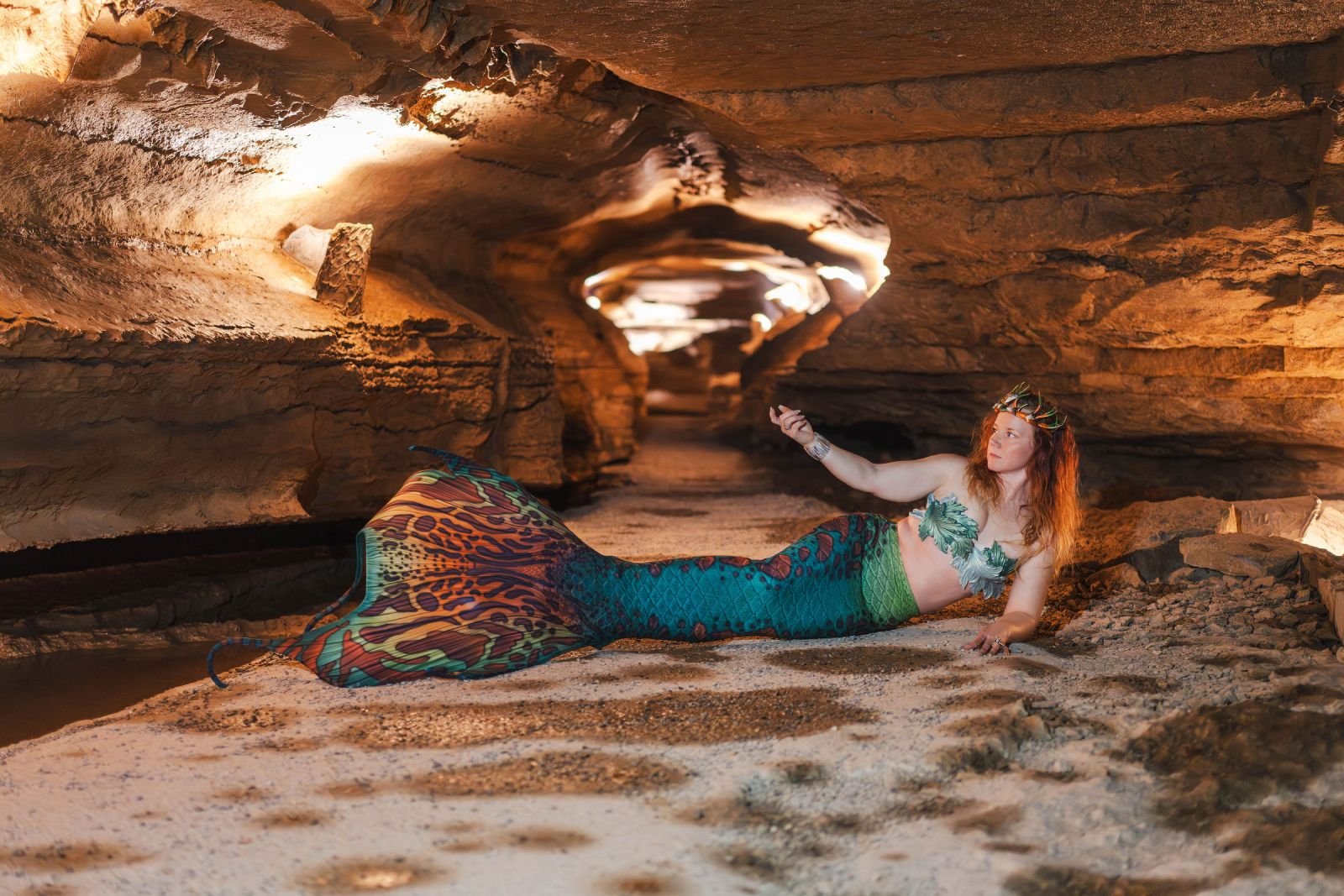 Crowned mermaid performer with a colorful turquoise tail reclining inside a warmly lit cave.
