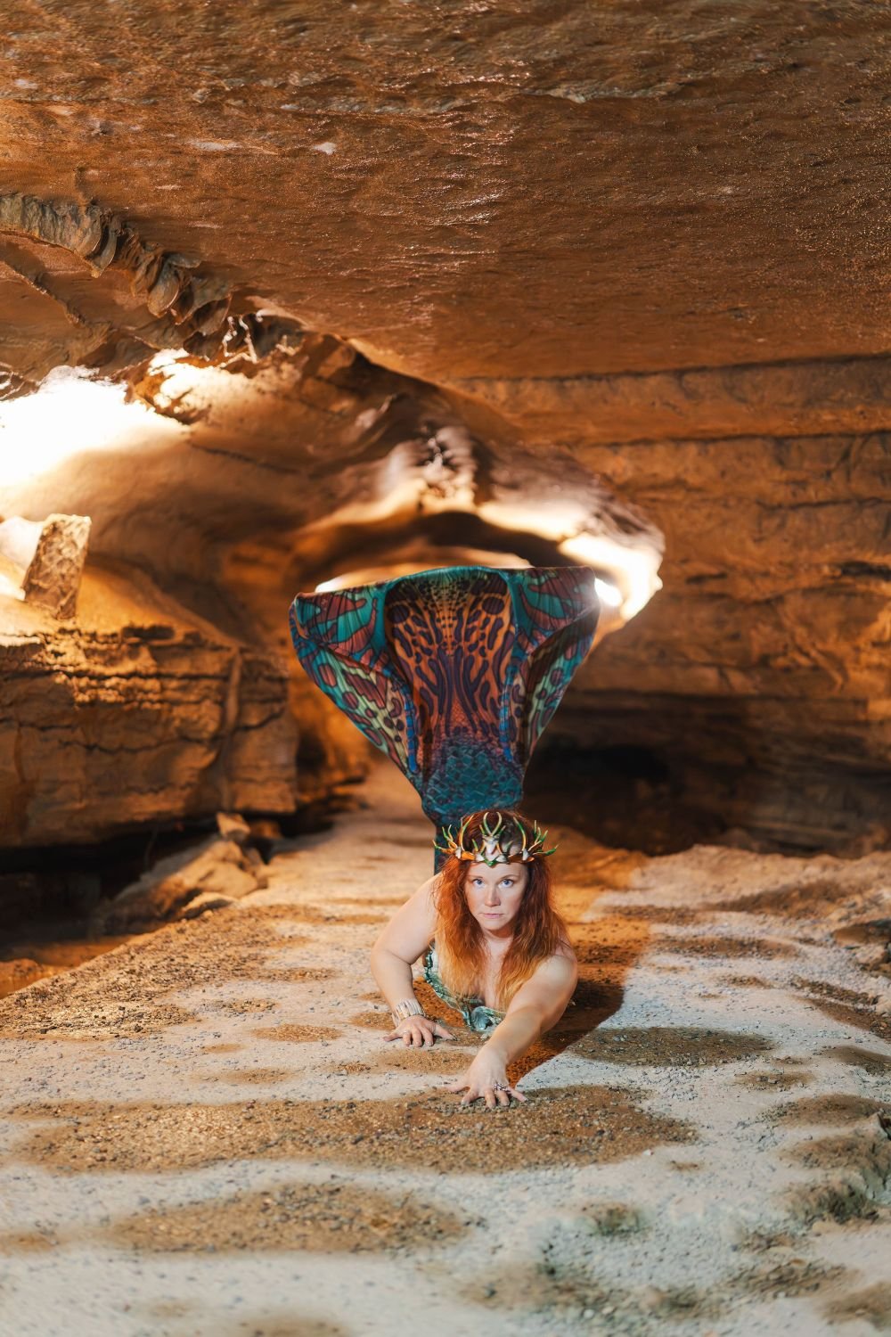 Crowned mermaid performer posing on a sandy cave floor with her colorful tail raised behind her.