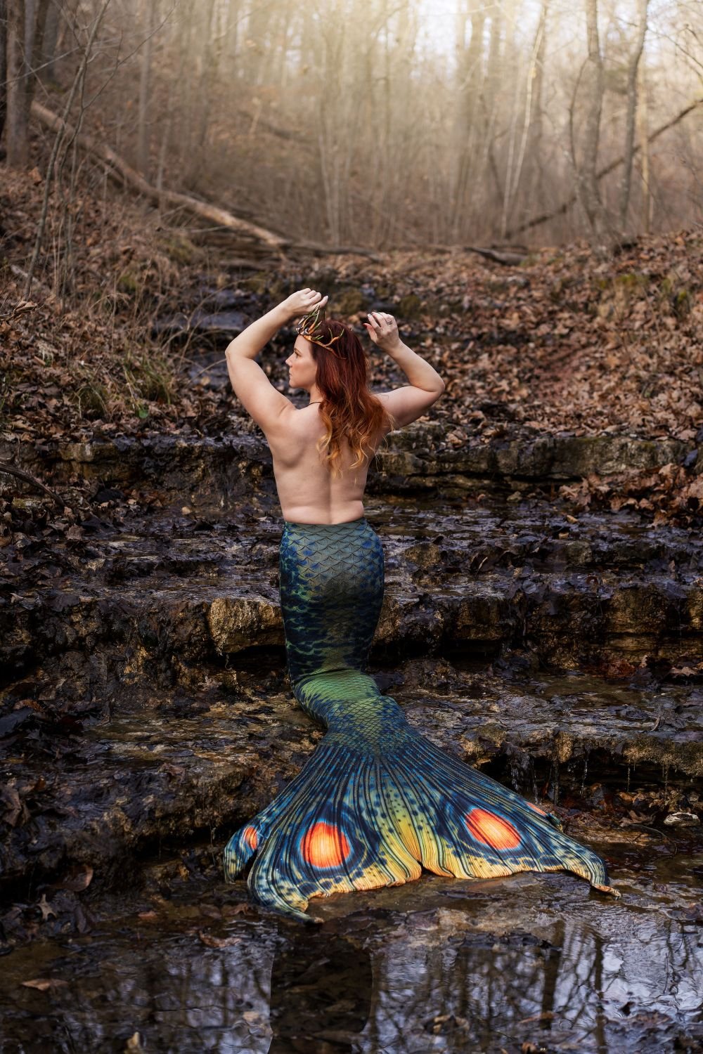 Mermaid performer viewed from behind wearing an antler crown and green patterned tail in a woodland stream.