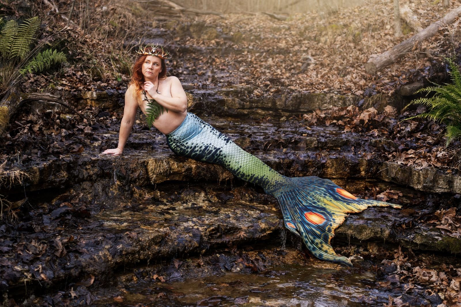 Mermaid performer wearing an antler crown and green patterned tail in a shallow woodland stream.
