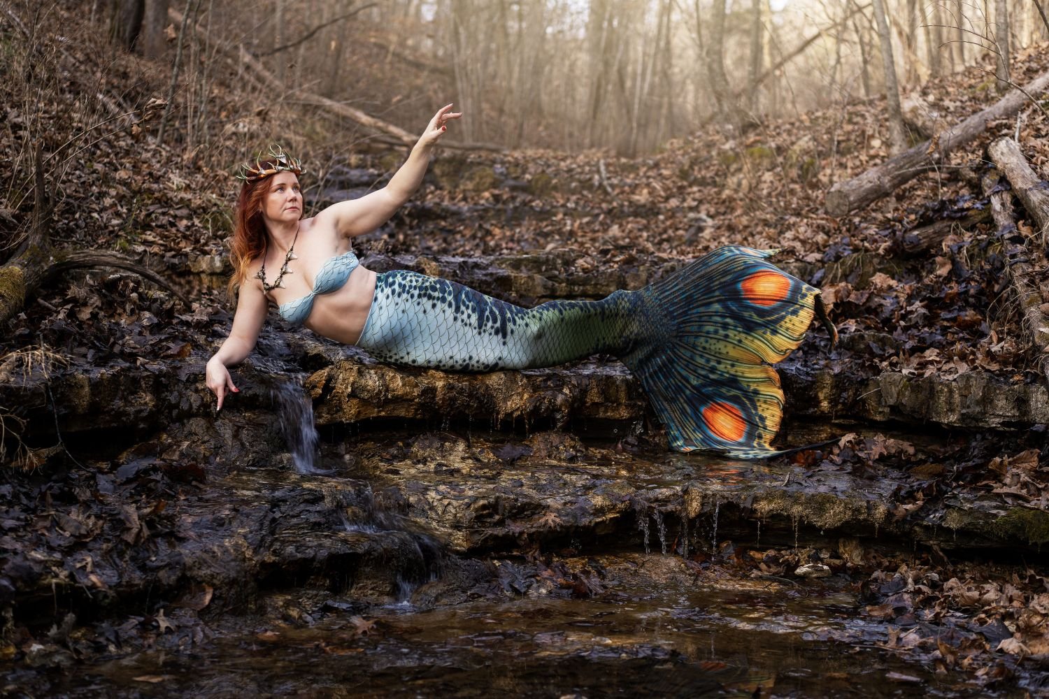 Mermaid performer wearing an antler crown and patterned tail beside a small woodland waterfall.