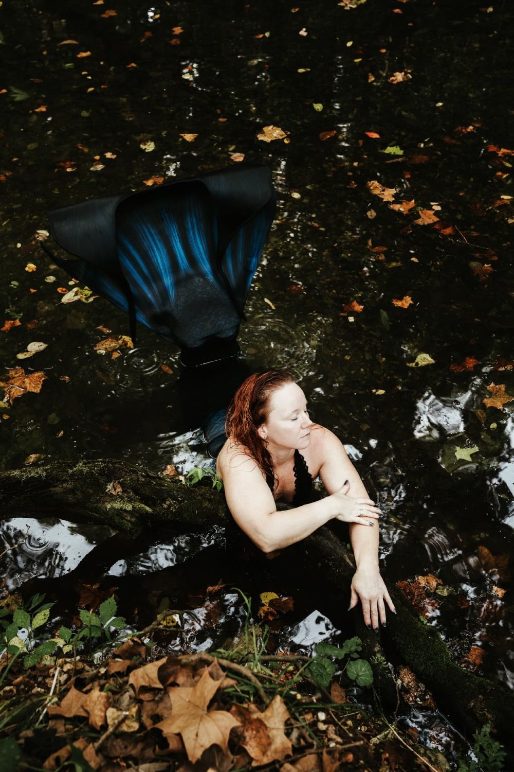 Mermaid performer with a dark blue tail resting against a mossy log in a woodland stream.