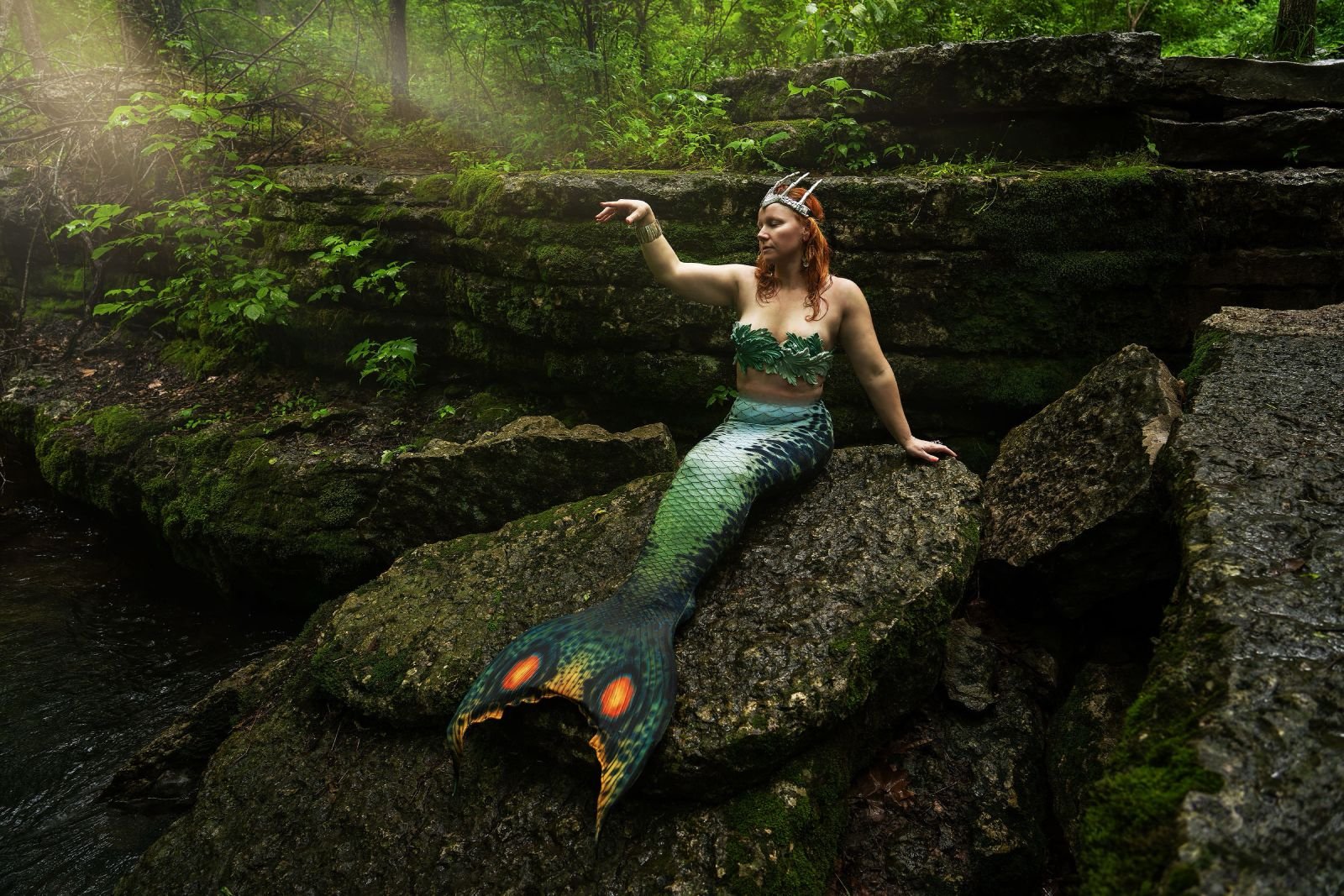Mermaid performer posed on moss-covered rocks beside a forest stream.