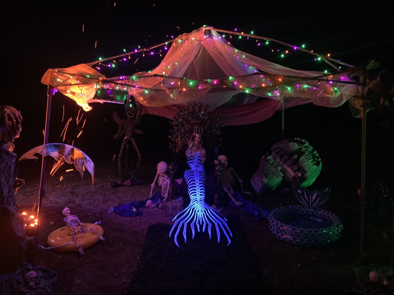Mermaid performer in a glowing skeleton tail surrounded by Halloween skeletons and colorful lights.