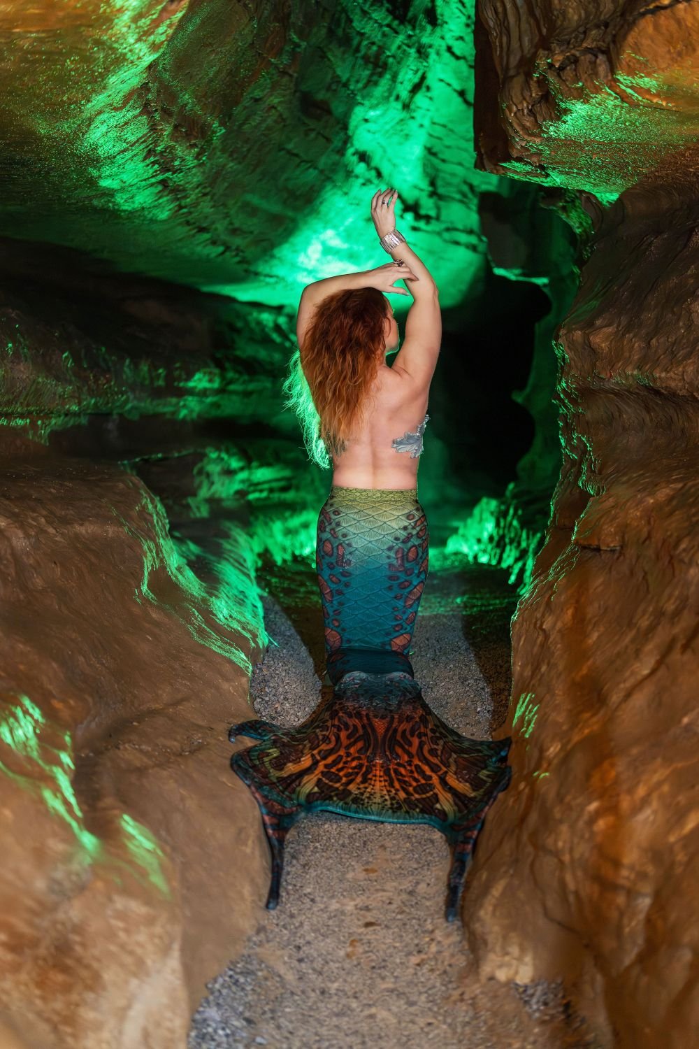 Mermaid performer with a patterned turquoise tail posing inside a cave illuminated by green light.
