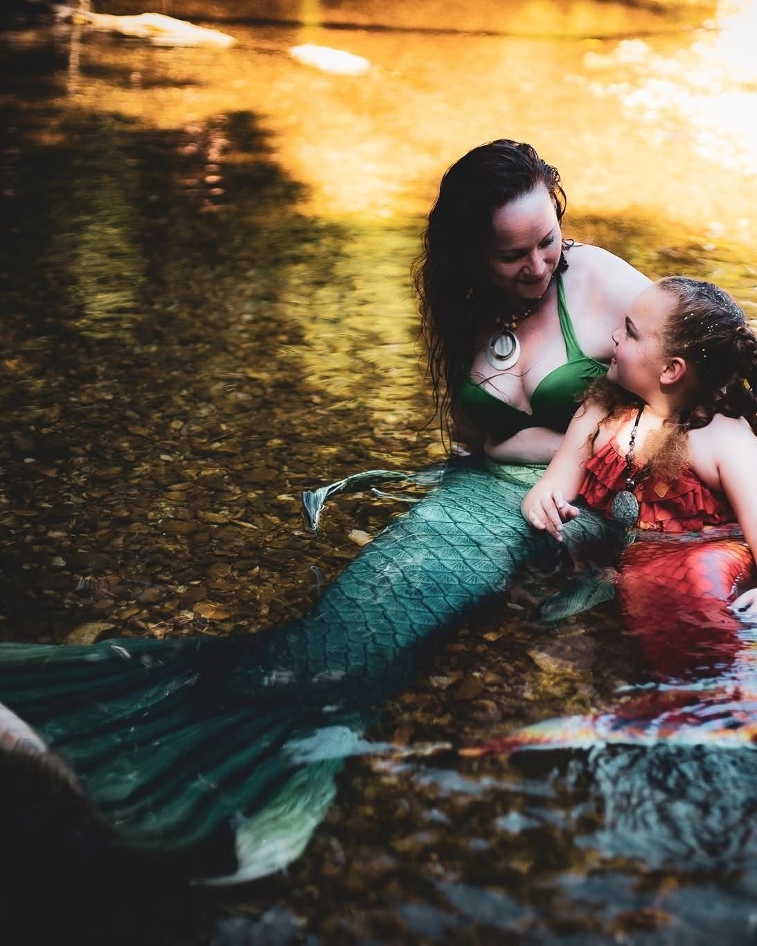 Mermaid performer and child in green and red tails smiling together in a shallow creek.