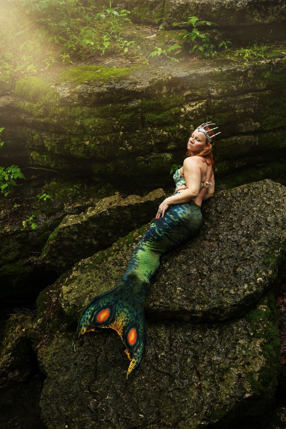 Mermaid performer with a green patterned tail reclining on moss-covered rocks in a forest.
