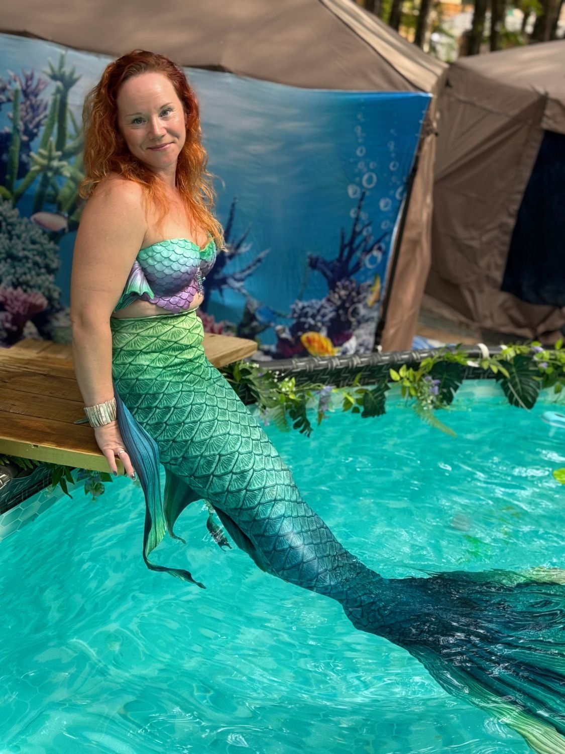 Mermaid performer with a green and blue tail seated beside a decorated outdoor pool.