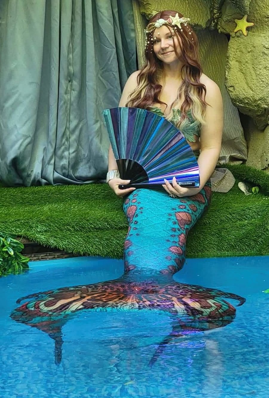 Mermaid performer with a turquoise patterned tail holding a blue fan beside a decorative pool.