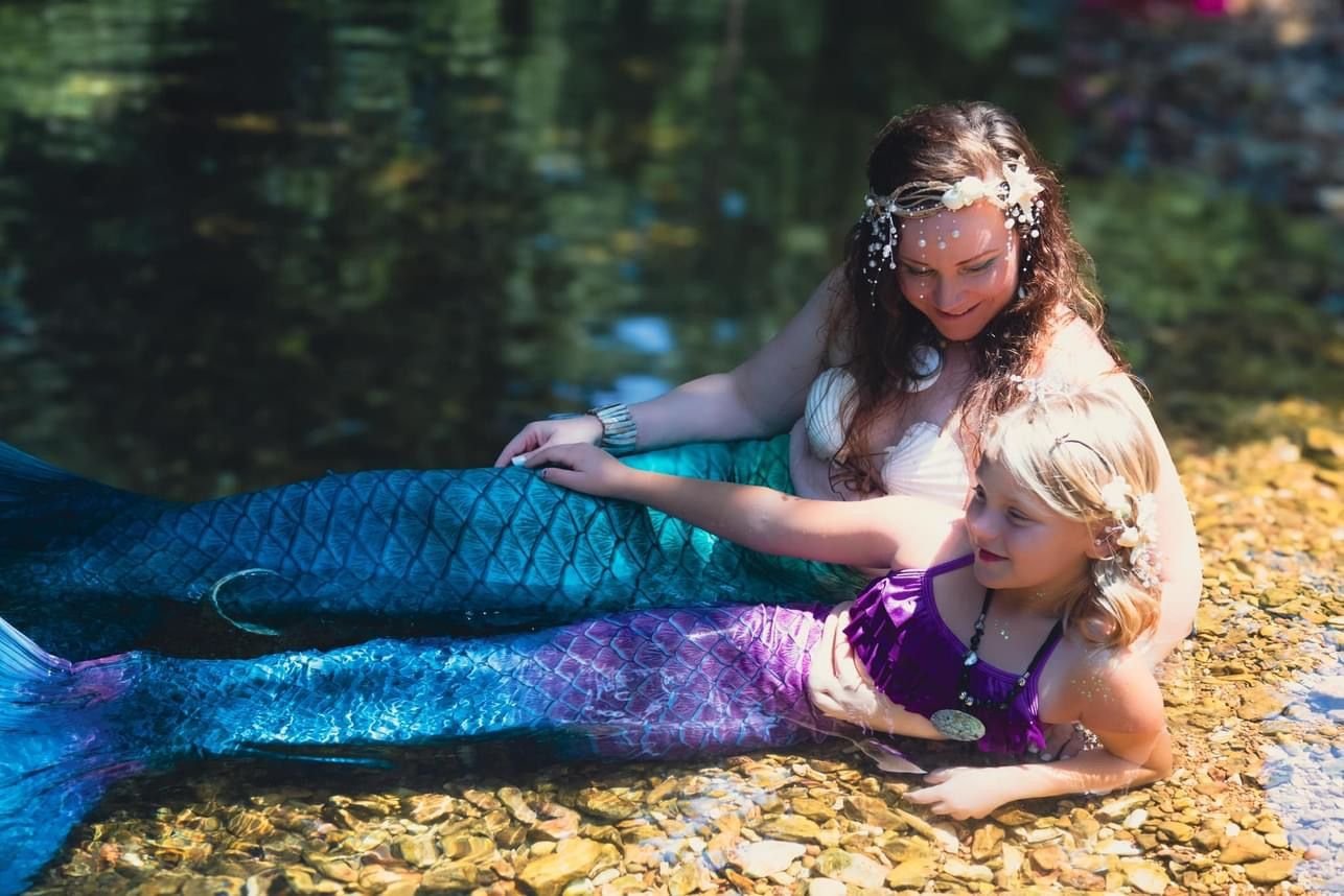 Mermaid performer and child in turquoise and purple tails relaxing together in a shallow creek.