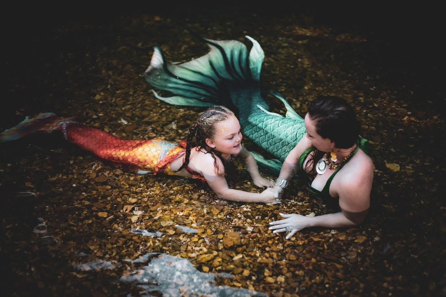 Mermaid performer and child in green and orange tails holding hands in a shallow woodland creek.