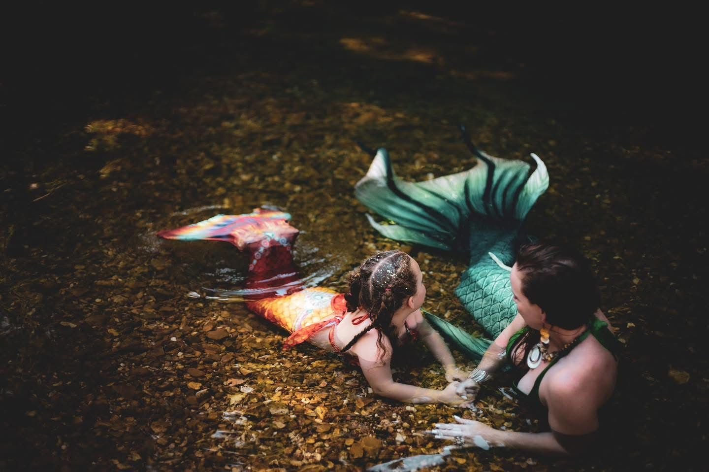 Mermaid performer and child in green and orange tails holding hands in a shallow creek.