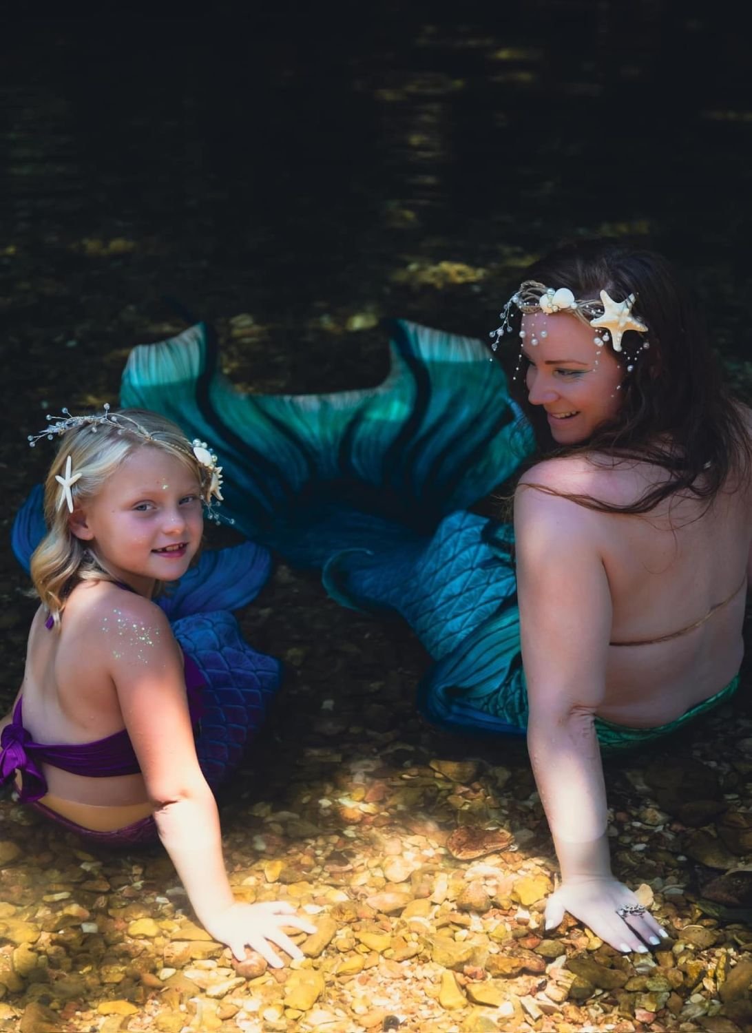Mermaid performer and child wearing colorful tails and seashell headpieces in a shallow woodland stream.