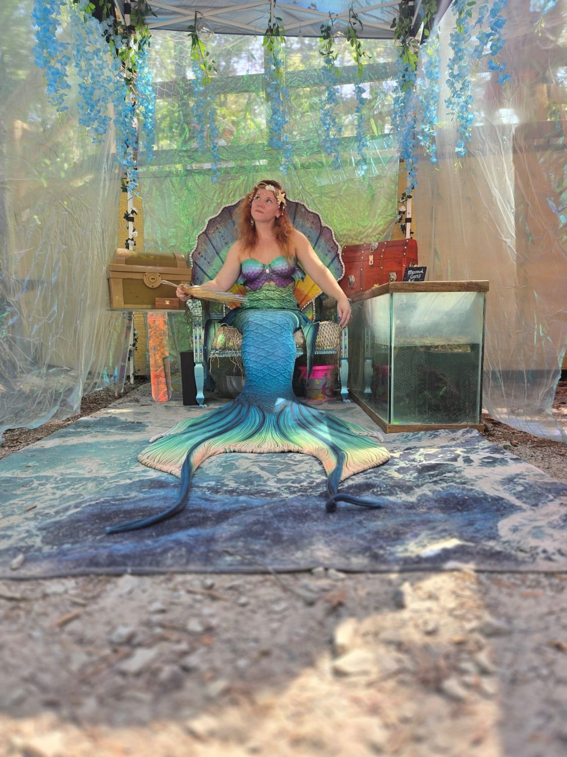 Mermaid performer with a blue-green tail seated among flowers, treasure chests, and ocean-themed decorations.