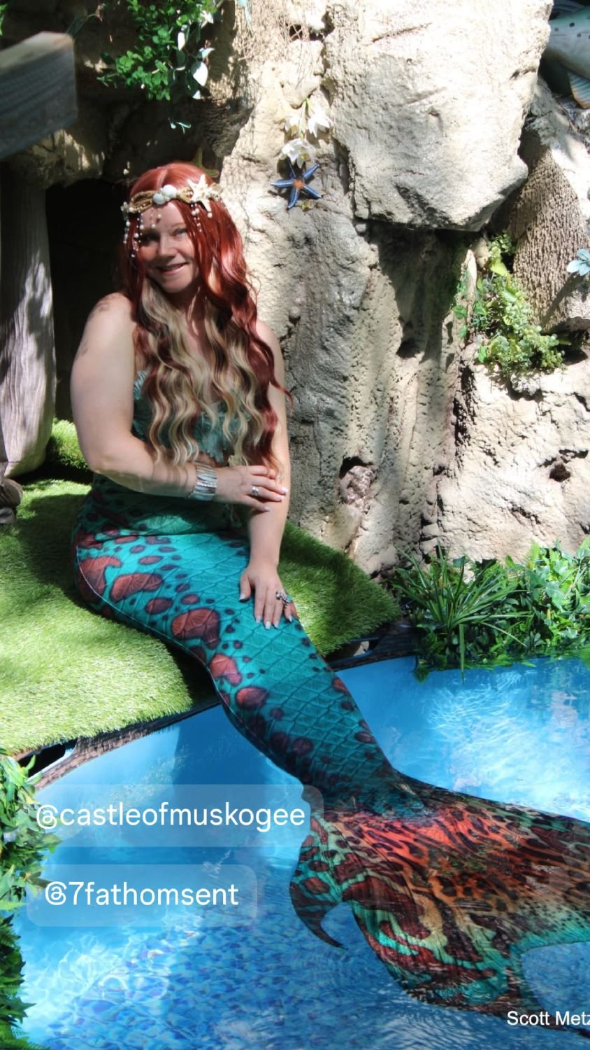 Mermaid performer with a colorful turquoise tail seated beside a decorative water display.