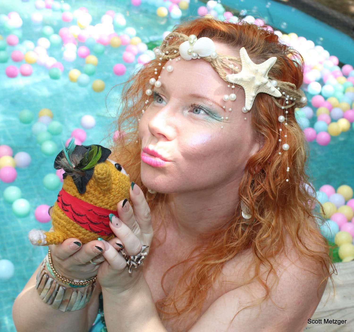 Mermaid performer wearing a seashell headpiece holding a handmade toy beside a pastel-filled pool.