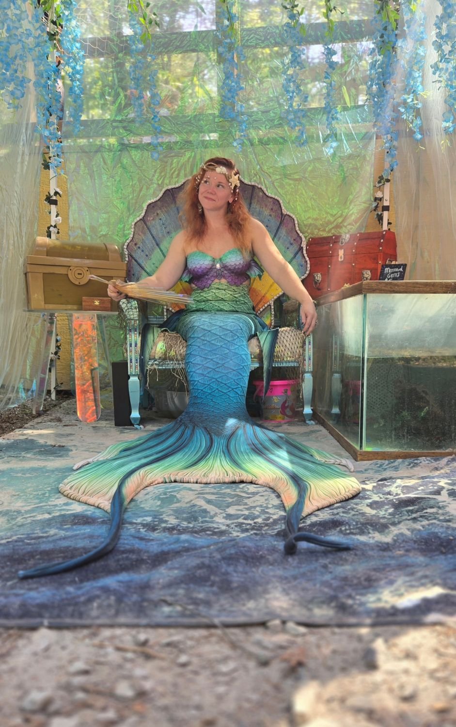 Mermaid performer with a blue-green tail seated on a throne beside treasure chests and ocean-themed decorations.