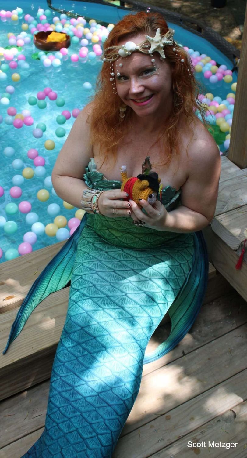 Mermaid performer in a turquoise tail holding a small toy beside a pool filled with pastel balls.