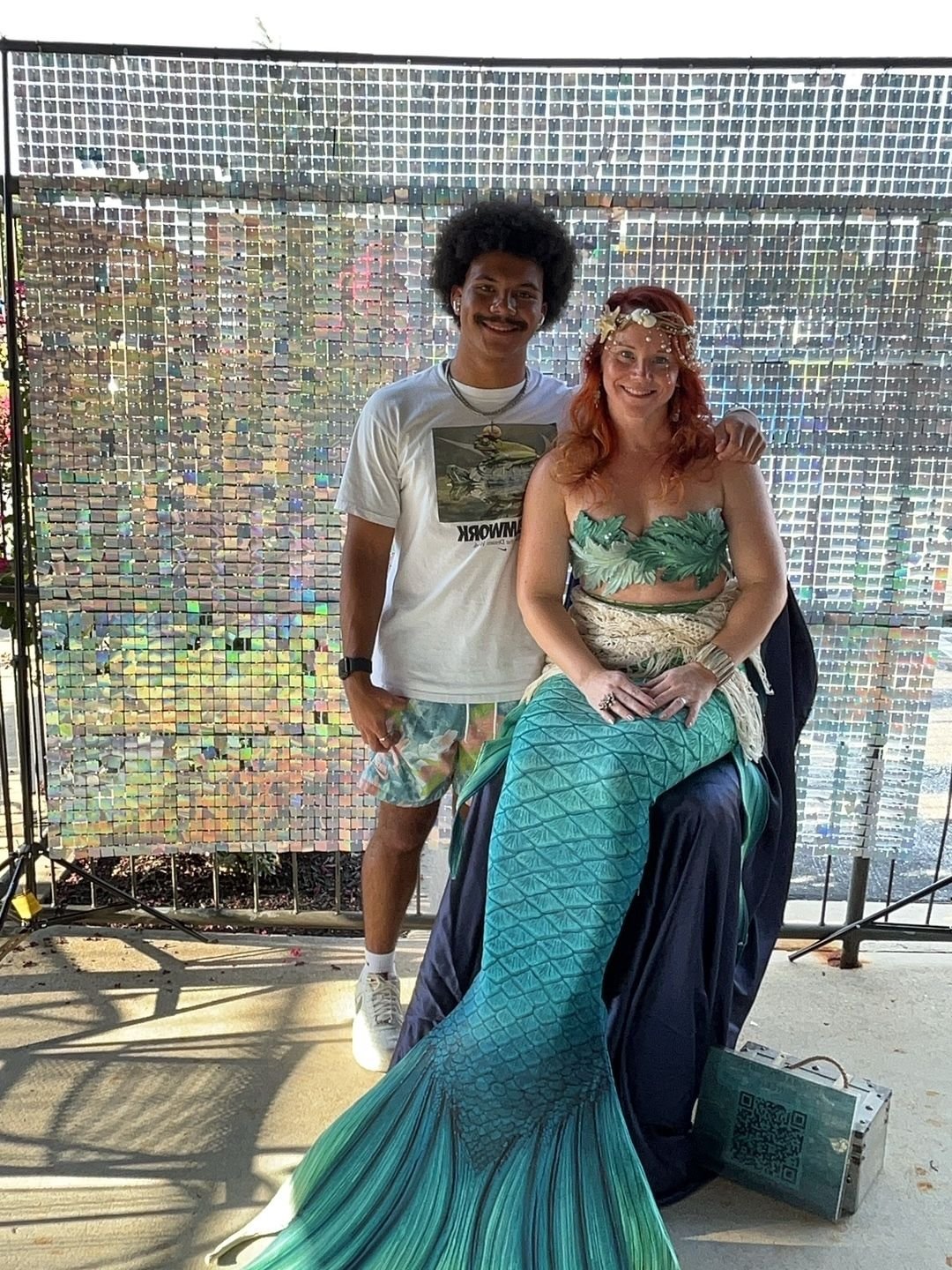 Mermaid performer with a turquoise tail posing beside a guest against a shimmering backdrop.