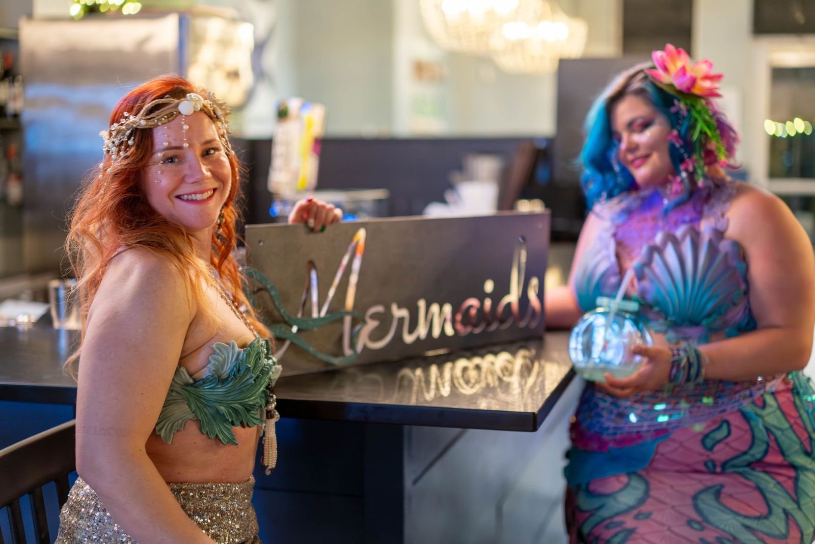Two mermaid performers in ocean-themed costumes posing beside a decorative Mermaids sign at an indoor bar.