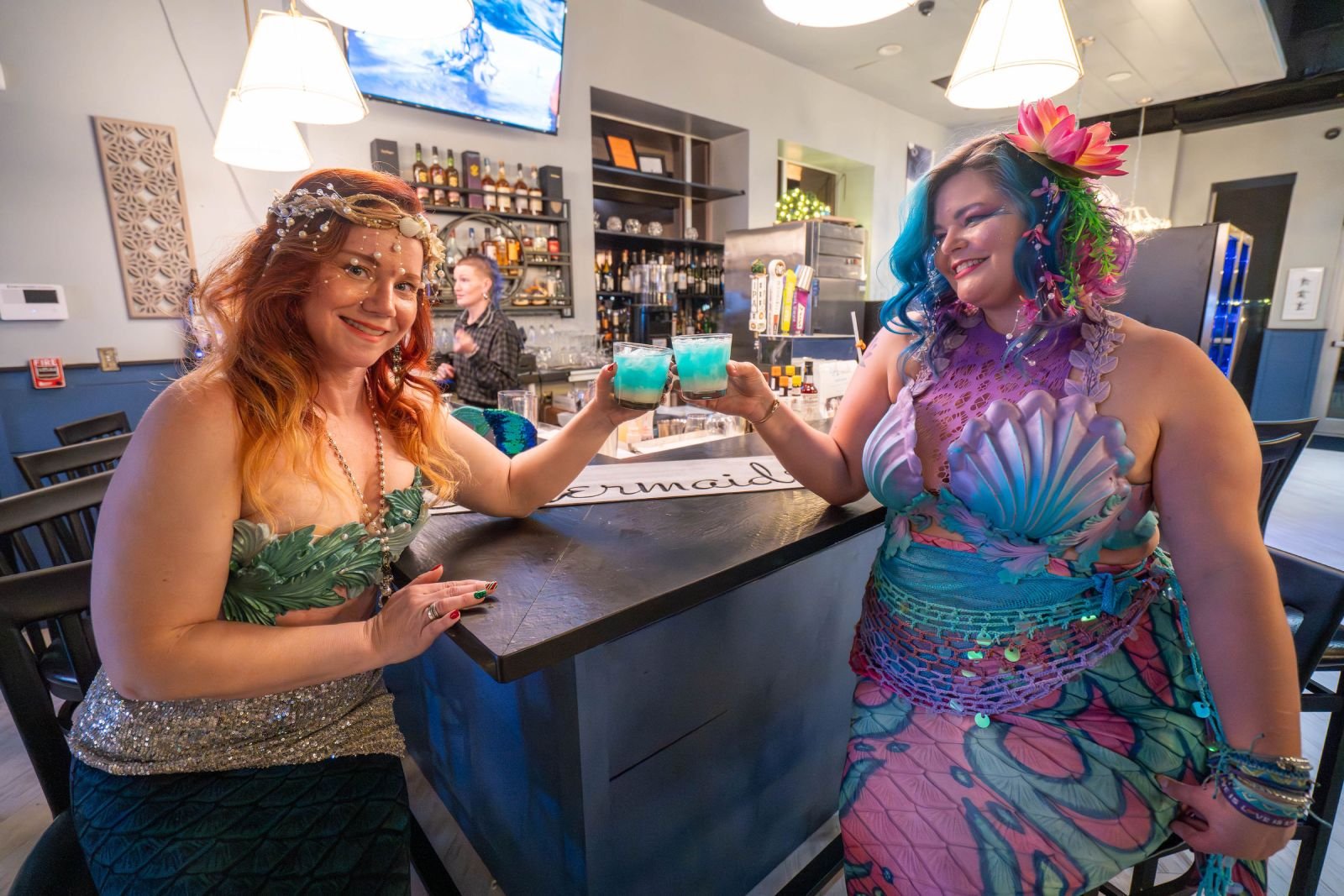 Two mermaid performers in colorful costumes toast with blue drinks at an indoor bar.