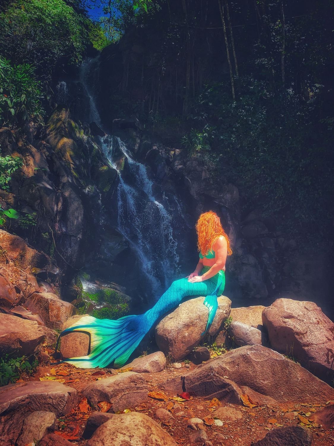 Professional mermaid performer with a turquoise tail seated on rocks beside a forest waterfall.
