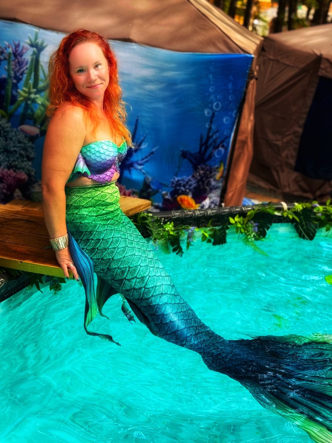 Professional mermaid performer with a green and blue tail posing beside a pool.