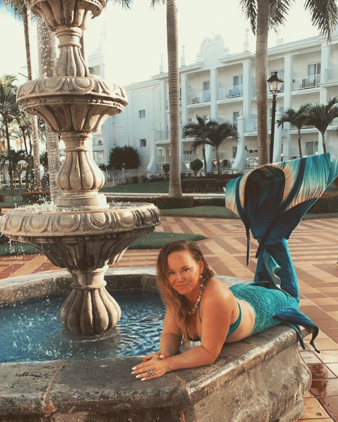 Mermaid performer with a turquoise tail reclining beside an ornate fountain in a palm-lined courtyard.
