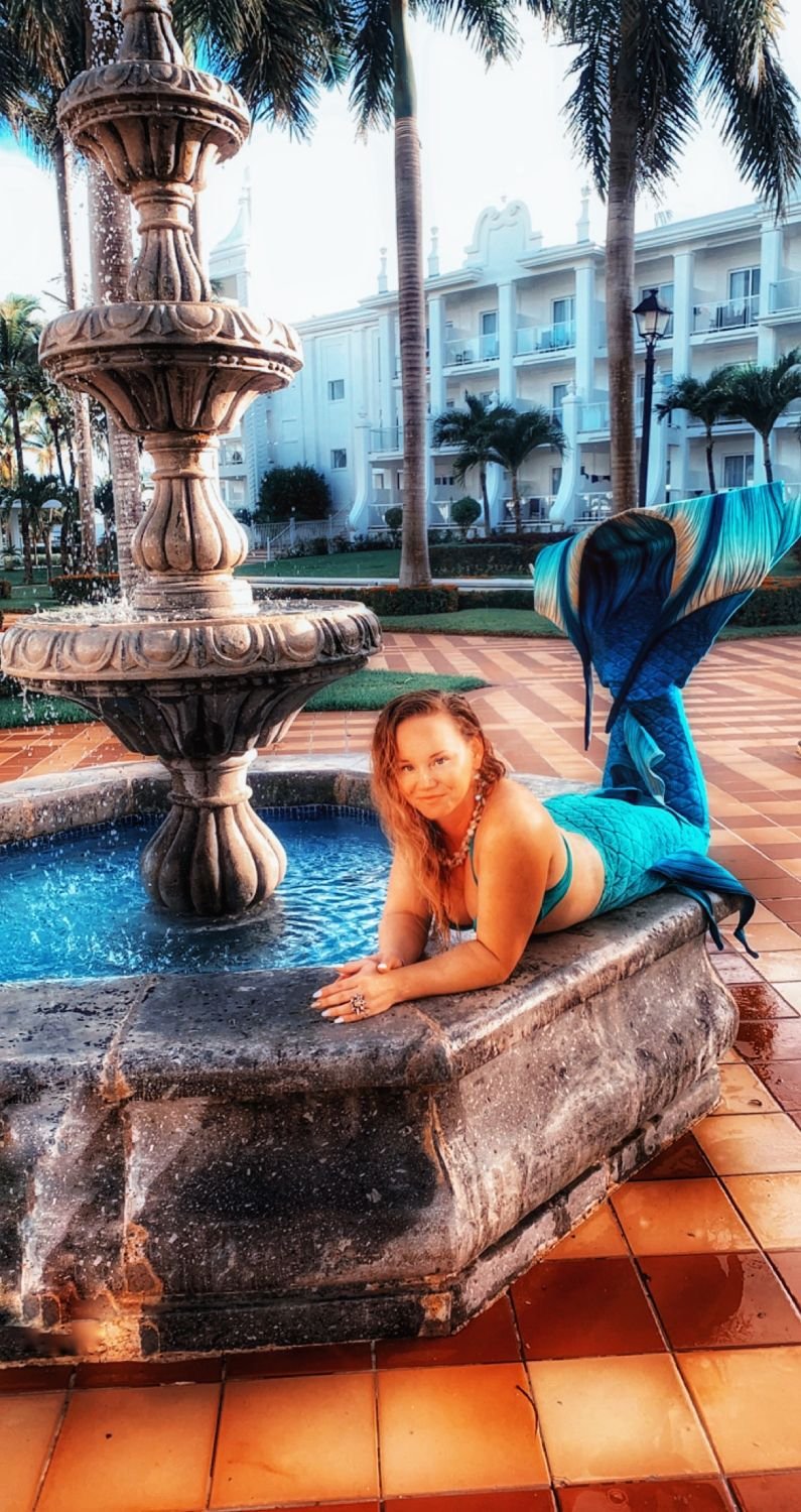 Mermaid performer with a turquoise tail posing beside an ornate courtyard fountain.