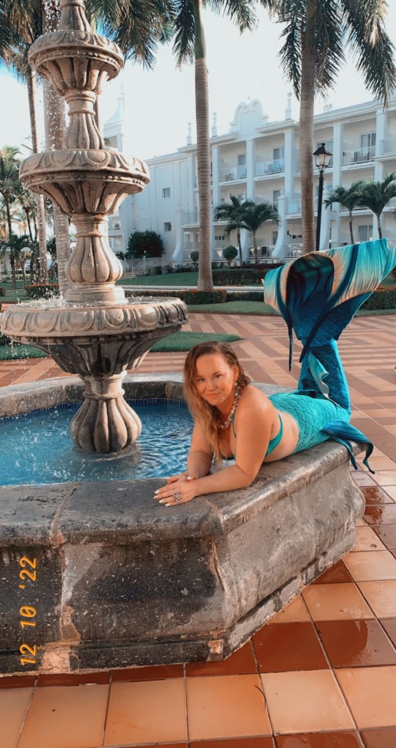 Mermaid performer with a turquoise tail reclining beside an ornate fountain in a tropical courtyard.