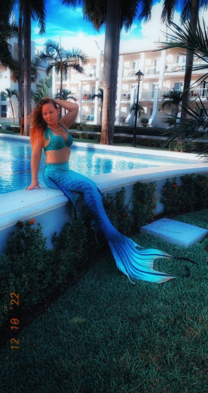 Mermaid performer with a turquoise tail seated beside a tropical resort pool.