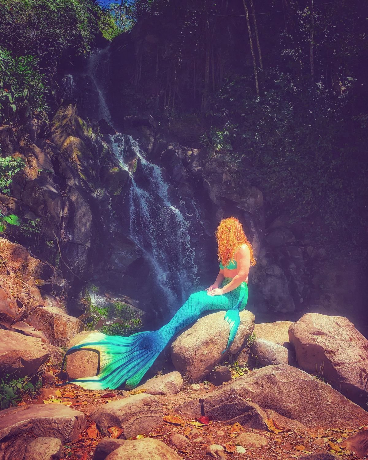 Mermaid performer with a turquoise tail resting on sunlit rocks beneath a forest waterfall.