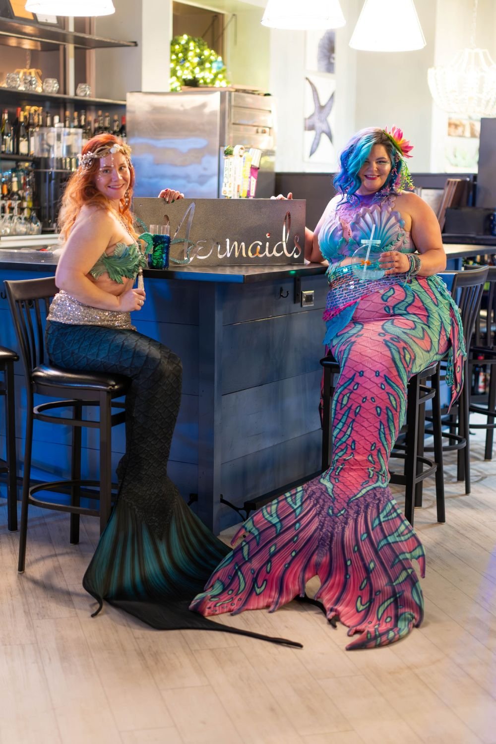 Two mermaid performers with green and pink tails holding drinks beside a decorative Mermaids sign.