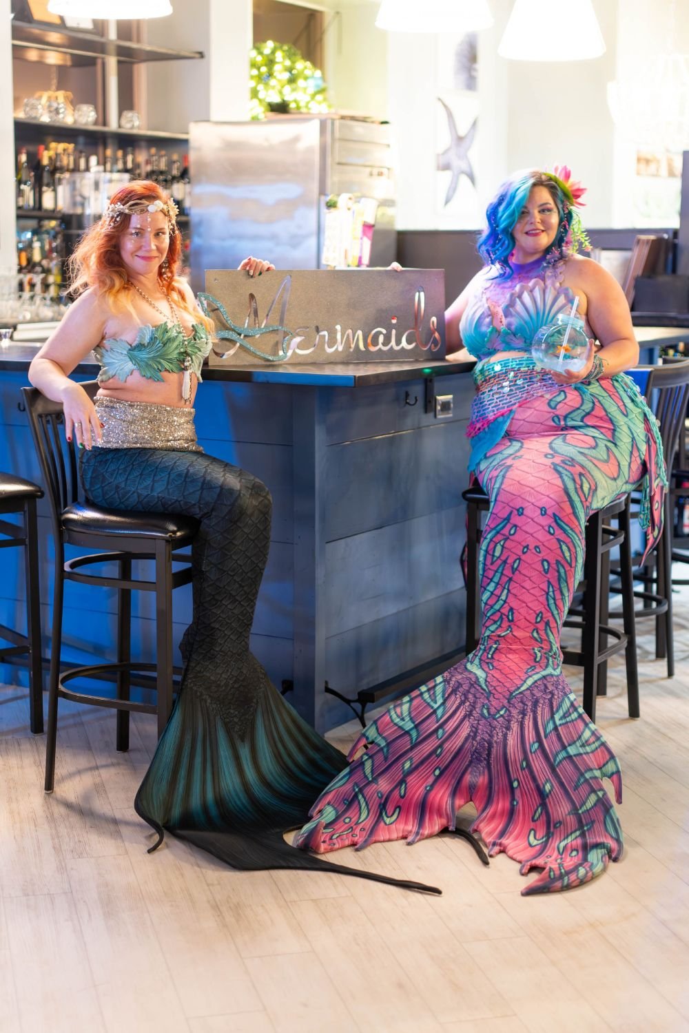 Two mermaid performers in green and pink tails seated together at an indoor bar.