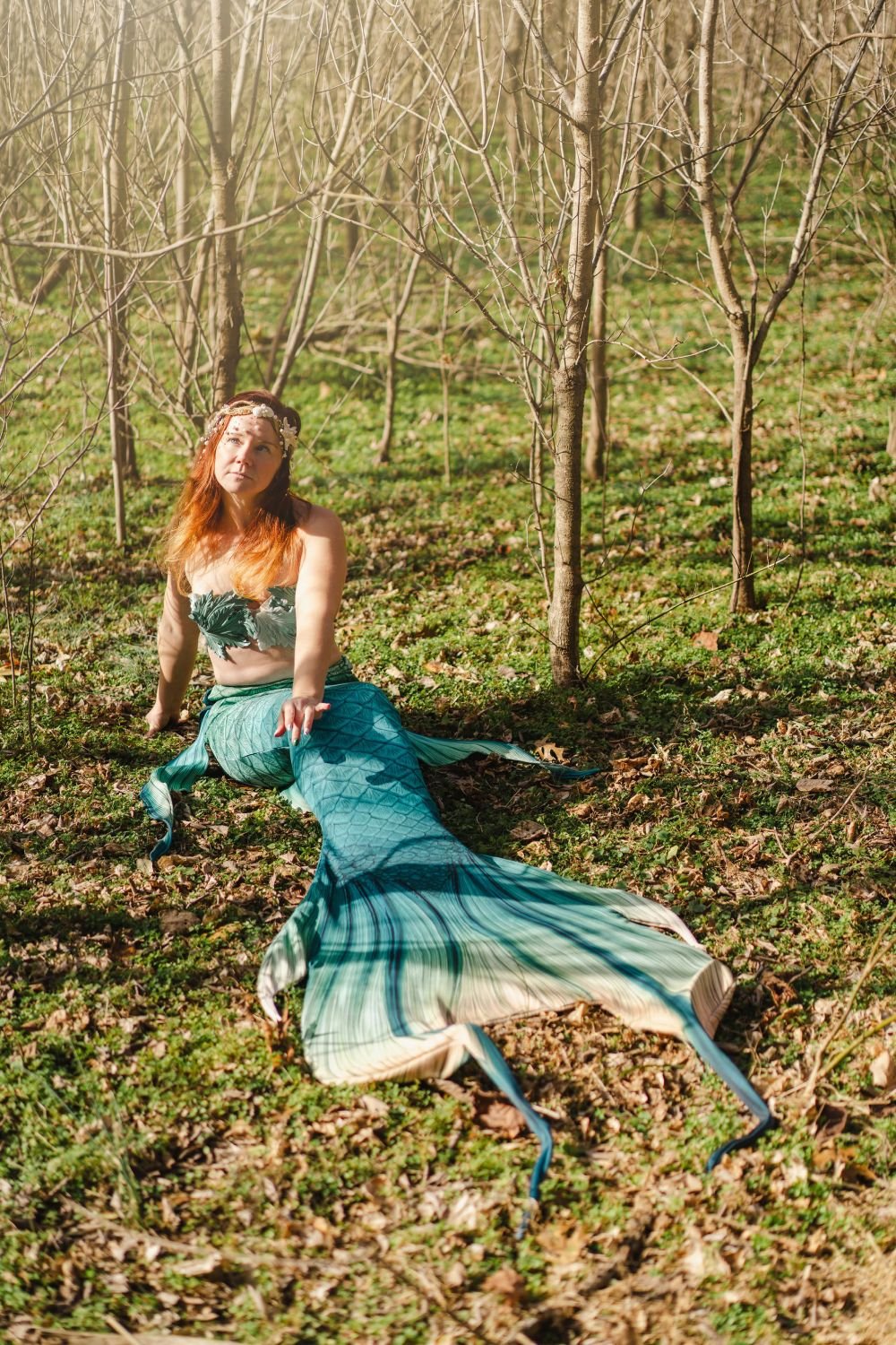 Mermaid performer with a flowing green tail seated among young trees on a woodland floor.