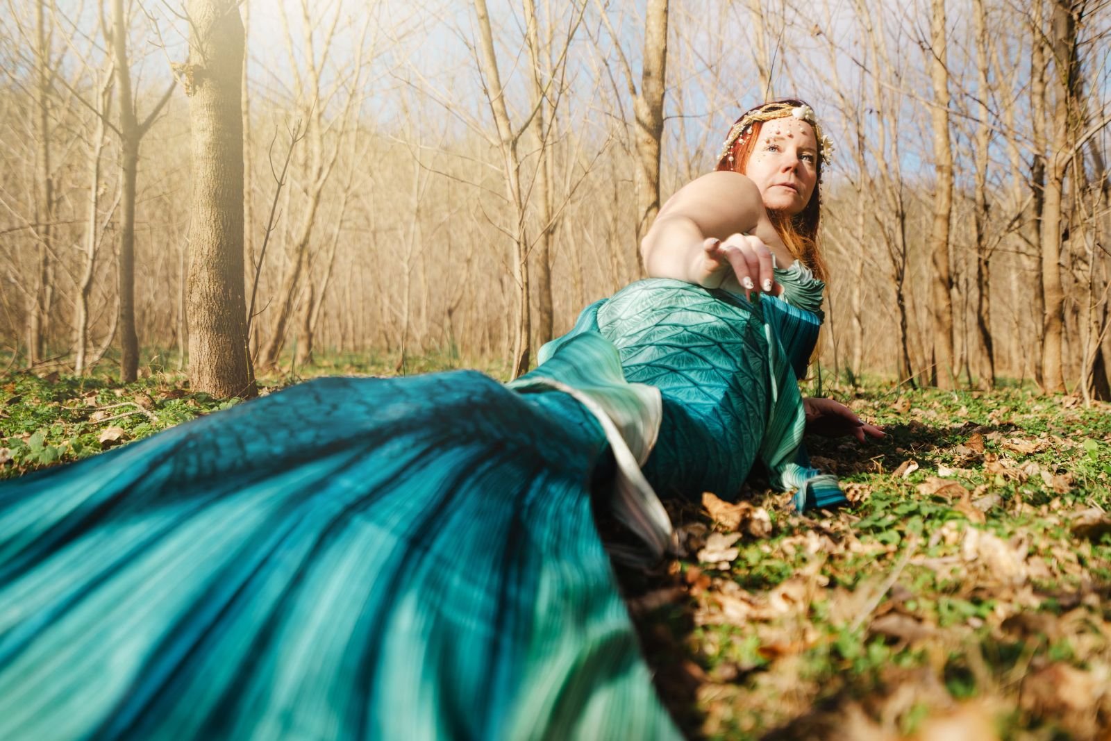 Mermaid performer with a turquoise tail reaching forward while reclining on a sunlit woodland floor.