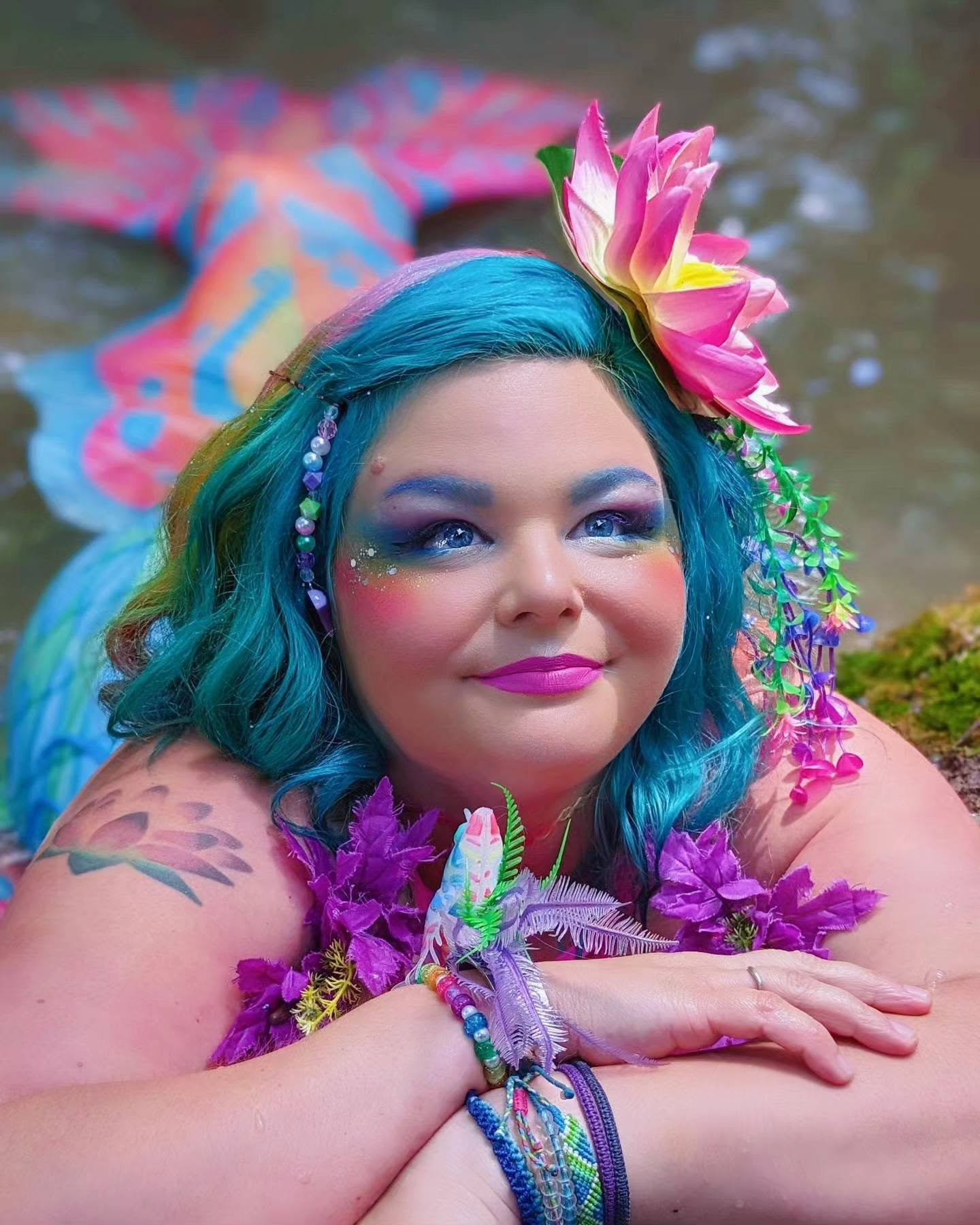 WhimSea Waves smiling in a colorful mermaid look with bright hair, flower accessories, and a rainbow tail in the background.