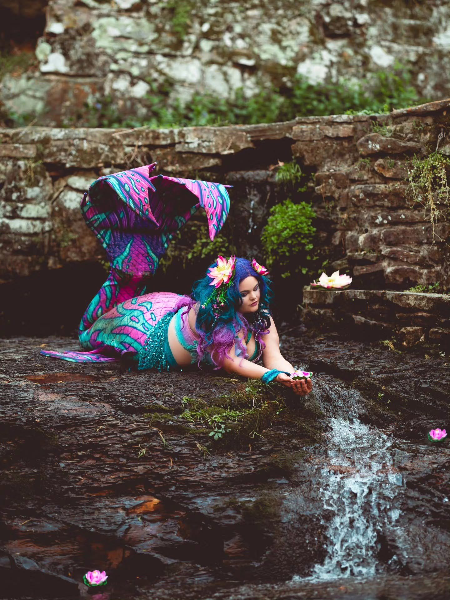 WhimSea Waves resting near a rocky waterfall in a colorful rainbow mermaid tail with flowers and fantasy styling.